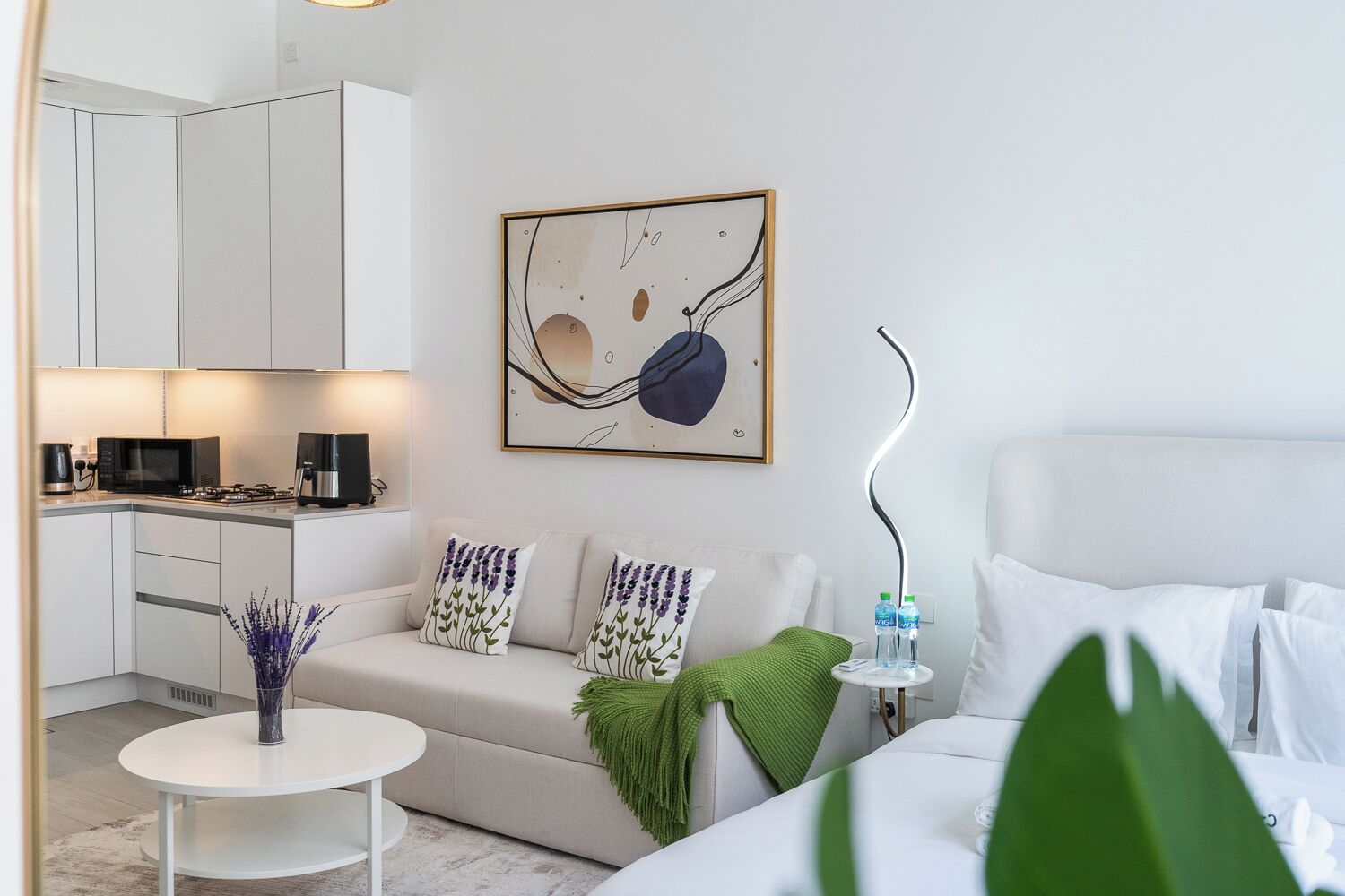 A modern hotel room with a kitchenette, a sofa with decorative pillows, a bedside table with water bottles, and a large abstract painting on the wall.