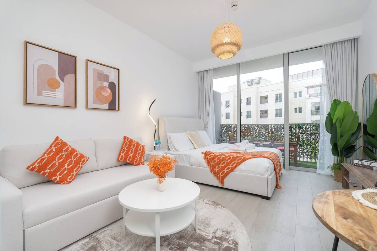 A modern hotel room with a white sofa, a bed with orange bedding, a round white coffee table, and a balcony with a view of buildings.