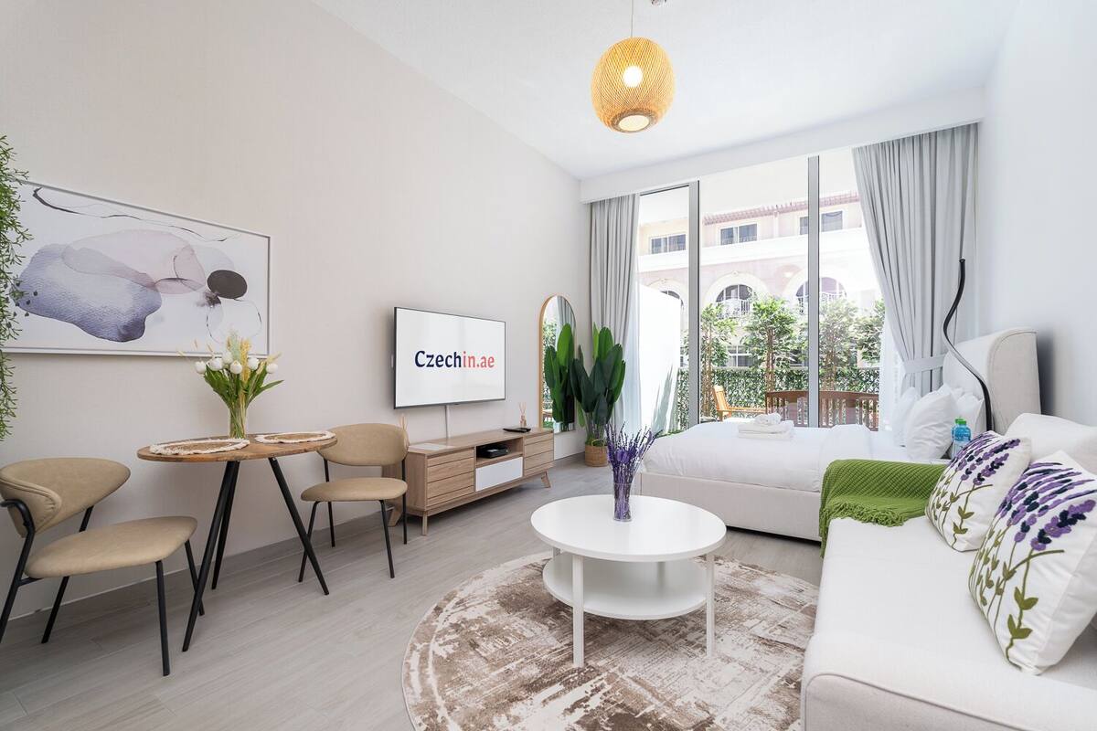 A modern hotel room with a bed, a TV, a round coffee table, and a dining area with chairs and a small table.