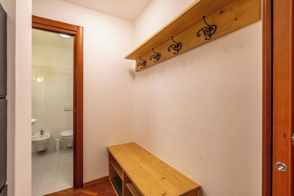 Un pasillo estrecho con un banco de madera, un estante de madera con ganchos y una puerta que da a un baño con inodoro y lavabo.
