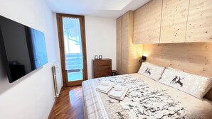 Un dormitorio con cama, cabecero de madera, cómoda, televisor montado en la pared y ventana con vistas.