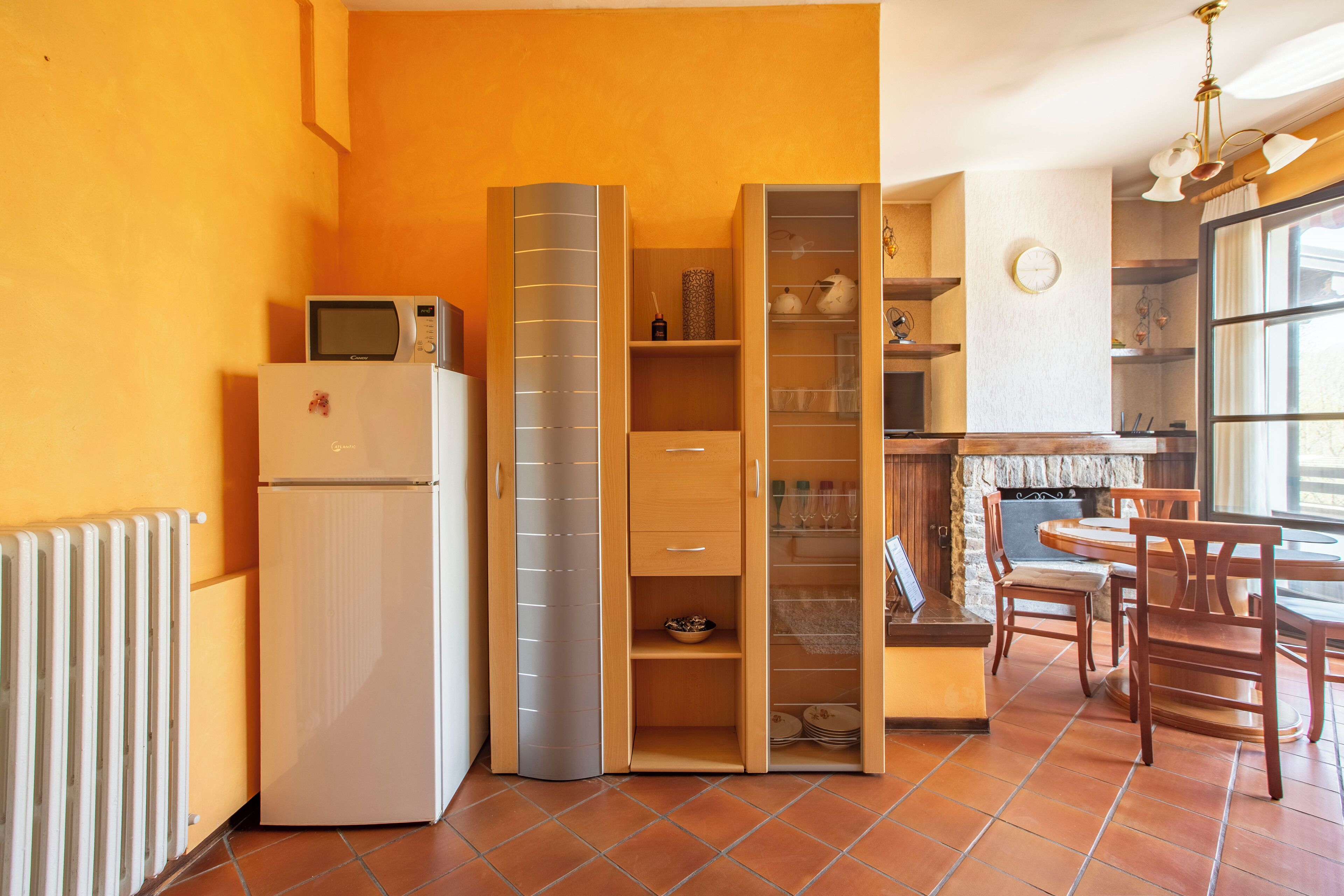 Een compacte keuken met een witte koelkast, een magnetron en een inbouwoven. Er is een houten kast met meerdere planken en een eethoek met een tafel en stoelen.