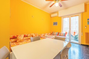 A room with a white table, a sofa with floral cushions, a ceiling fan, and a large window with curtains.