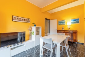 A room with a flat-screen TV, a dining table with chairs, a cabinet with a plant, and artwork on the walls.