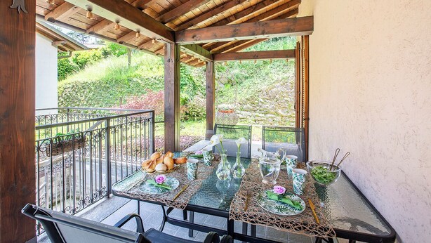 Un espacio de comedor al aire libre cubierto, con una mesa dispuesta para dos personas, rodeado por una pérgola de madera y vistas a un jardín.