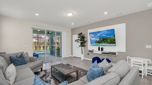 A modern living room with a grey sofa set, a coffee table, a flat-screen TV mounted on the wall, and a sliding glass door leading to a balcony.
