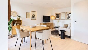 A modern living room with a wooden dining table, white chairs, a sofa, and a TV. There are framed pictures on the wall and a plant on the side. - The South Wimbledon Hideout - Fancy 1bdr Flat With Balcony (London)
