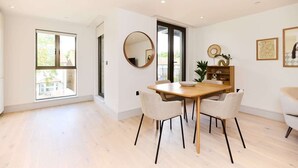 A modern dining area with a wooden table and four chairs, a round mirror on the wall, and a view of the outdoors through large windows. - The South Wimbledon Hideout - Fancy 1bdr Flat With Balcony (London)