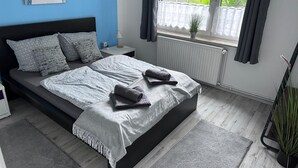 A modern bedroom with a large bed, two bedside tables, a nightstand, a window with curtains, a radiator, and a potted plant.