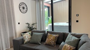 Ein modernes Wohnzimmer mit grauem Sofa, dekorativen Kissen, einer Wanduhr und einer Glastür, die zu einem Außenbereich führt.
