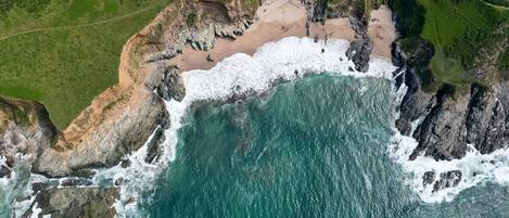 An aerial view of a coastal area with a beach, rocky cliffs, and a path leading to the beach.