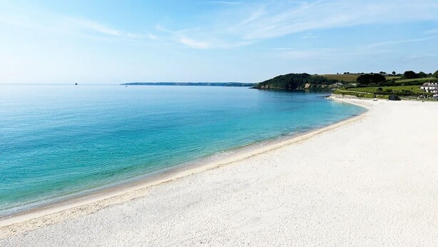 En bred sandstrand med klart blåt vand, en lille båd i det fjerne og et bølgende landskab med bygninger til højre.