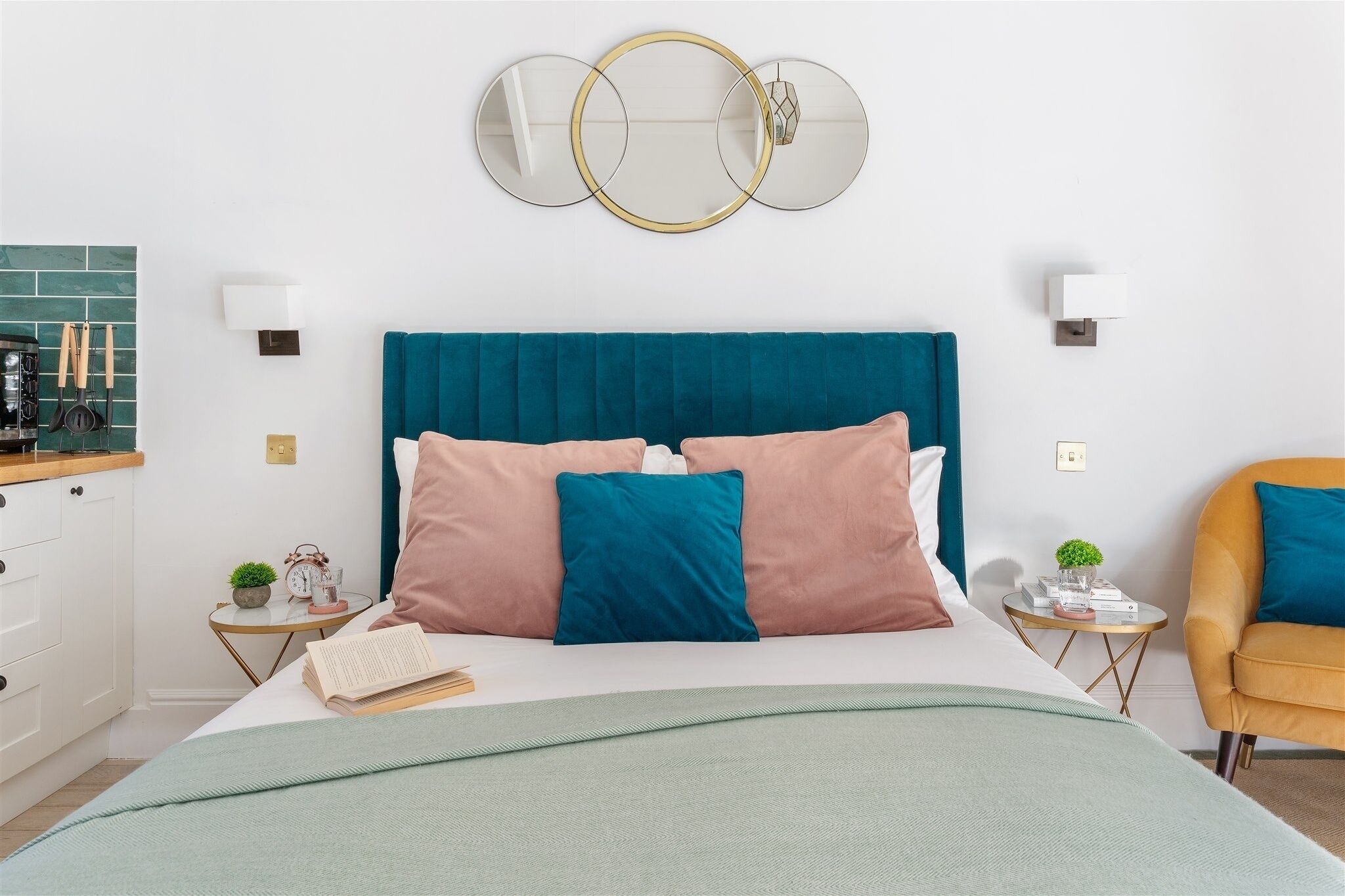 Une chambre à coucher bien rangée, avec une tête de lit turquoise, un lit agrémenté de coussins roses et turquoise, une table de chevet sur laquelle se trouvent un livre et une petite plante, et un fauteuil jaune.