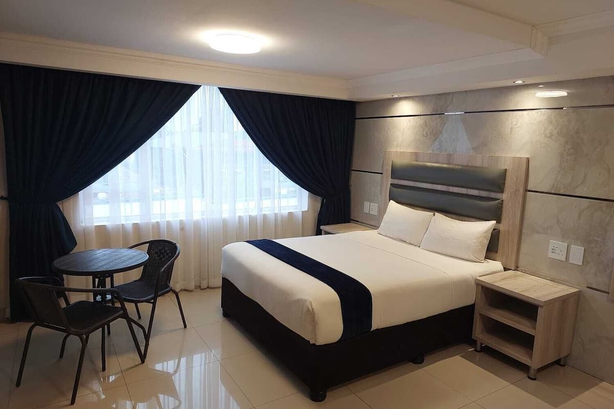 A hotel room with a bed, a small table, two chairs, a nightstand, and a view of the city through large windows.