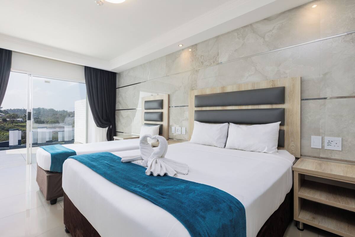 A modern hotel room with a large bed, a separate sofa bed, a balcony with curtains, and a view of the landscape.