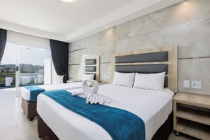 A modern hotel room with a large bed, a separate sofa bed, a balcony with curtains, and a view of the landscape. - Bayside Taj Hotel (Port Shepstone)