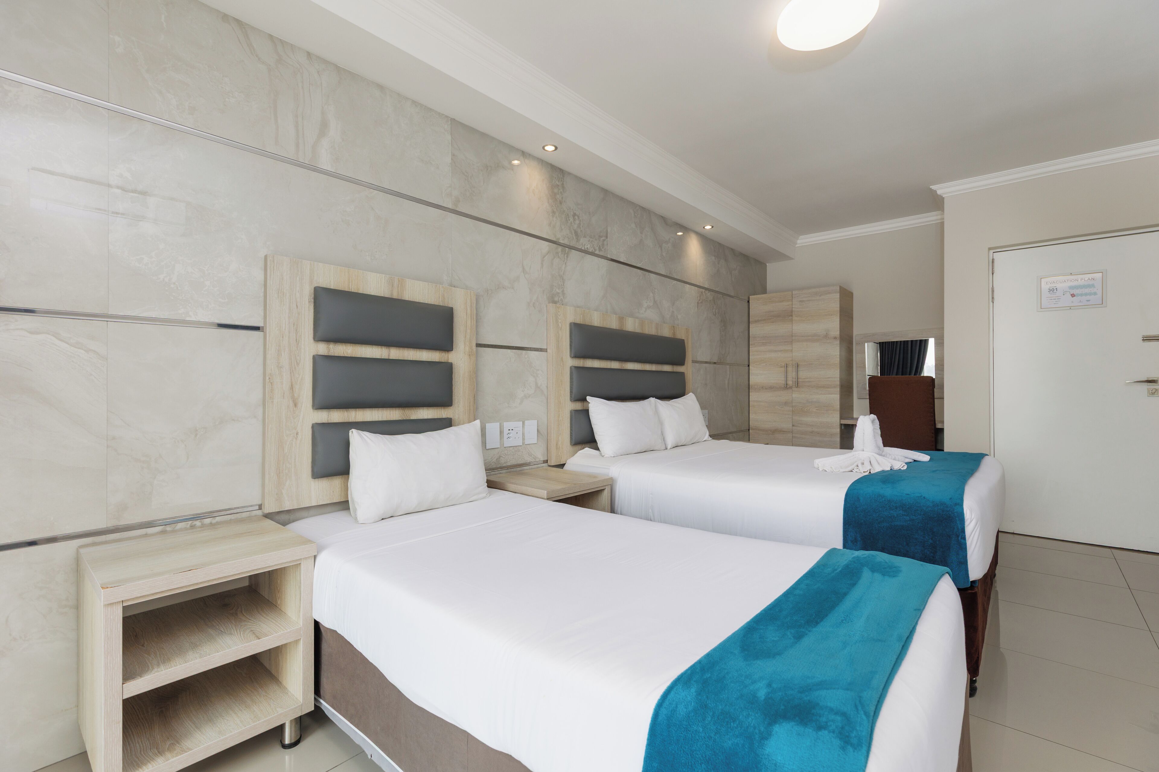A hotel room with two beds, a wooden headboard, a nightstand, and a small wooden shelf.