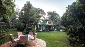 Um pátio com jardim, com mesa e cadeiras de tijolo, cercado por vegetação exuberante e uma casa ao fundo.