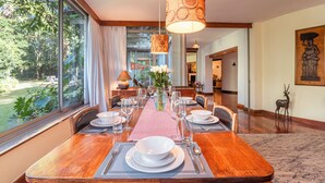 Uma área de refeições com uma mesa grande posta para uma refeição, rodeada por cadeiras, um vaso com flores e vista para o exterior.
