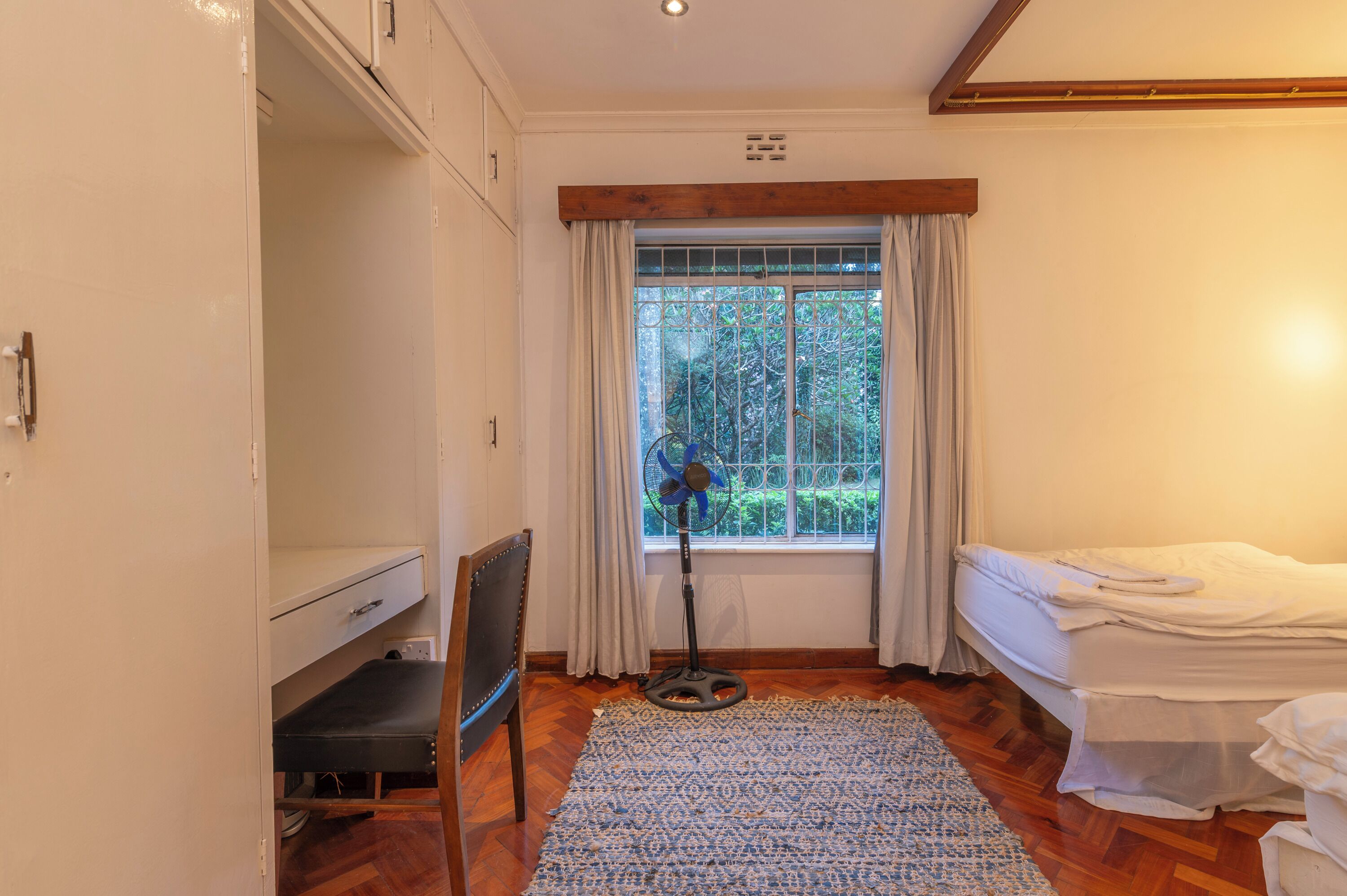 Une chambre à coucher comprenant un lit, un bureau, une chaise, un ventilateur sur pied et une fenêtre avec des rideaux.