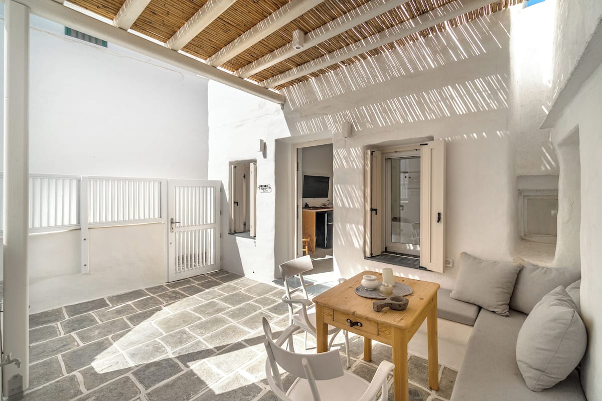 A sunlit patio with a stone floor, a wooden table, white chairs, a sofa, and a door leading to an interior space.