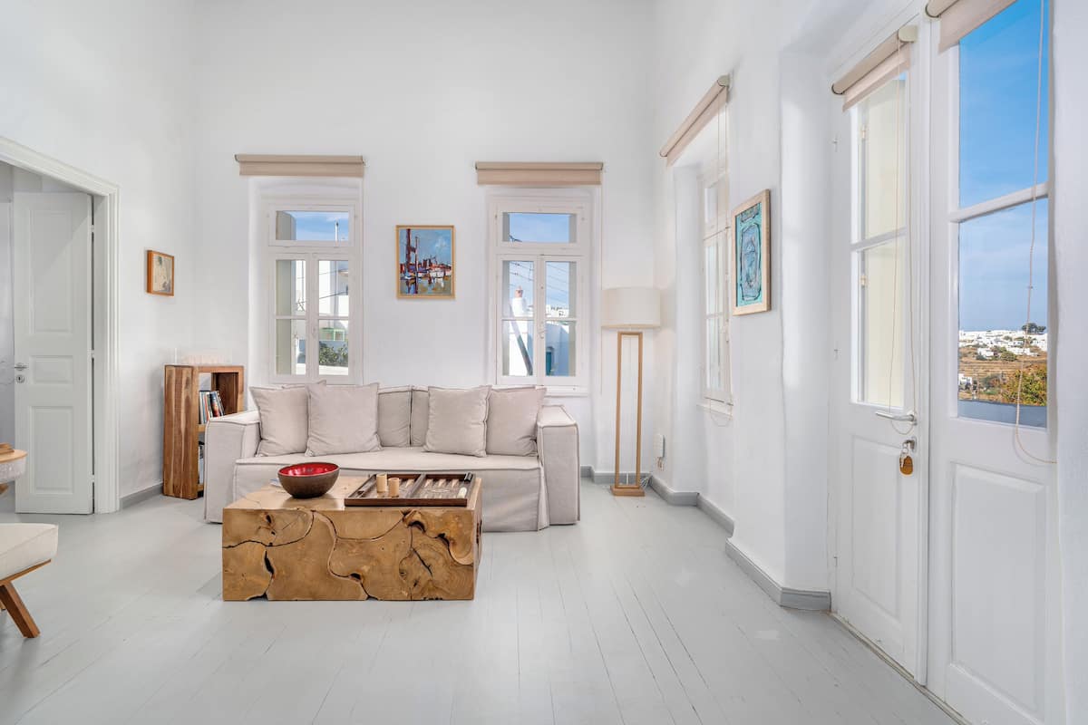A spacious living room with a white sofa, wooden coffee table, and large windows offering a view of the city.