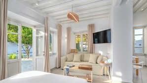 A modern hotel room with a bed, sofa, TV, and large windows offering a view of the outdoors. - Rabagas Suites (Sifnos)