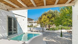 A covered outdoor area with a stone floor, a pool, and a view of a white building with a window. - Rabagas Suites (Sifnos)