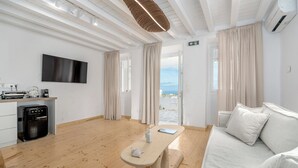A modern hotel room with a wooden floor, a sofa, a table with a bottle and a notebook, a TV on the wall, and a view of the sea through the window. - Rabagas Suites (Sifnos)