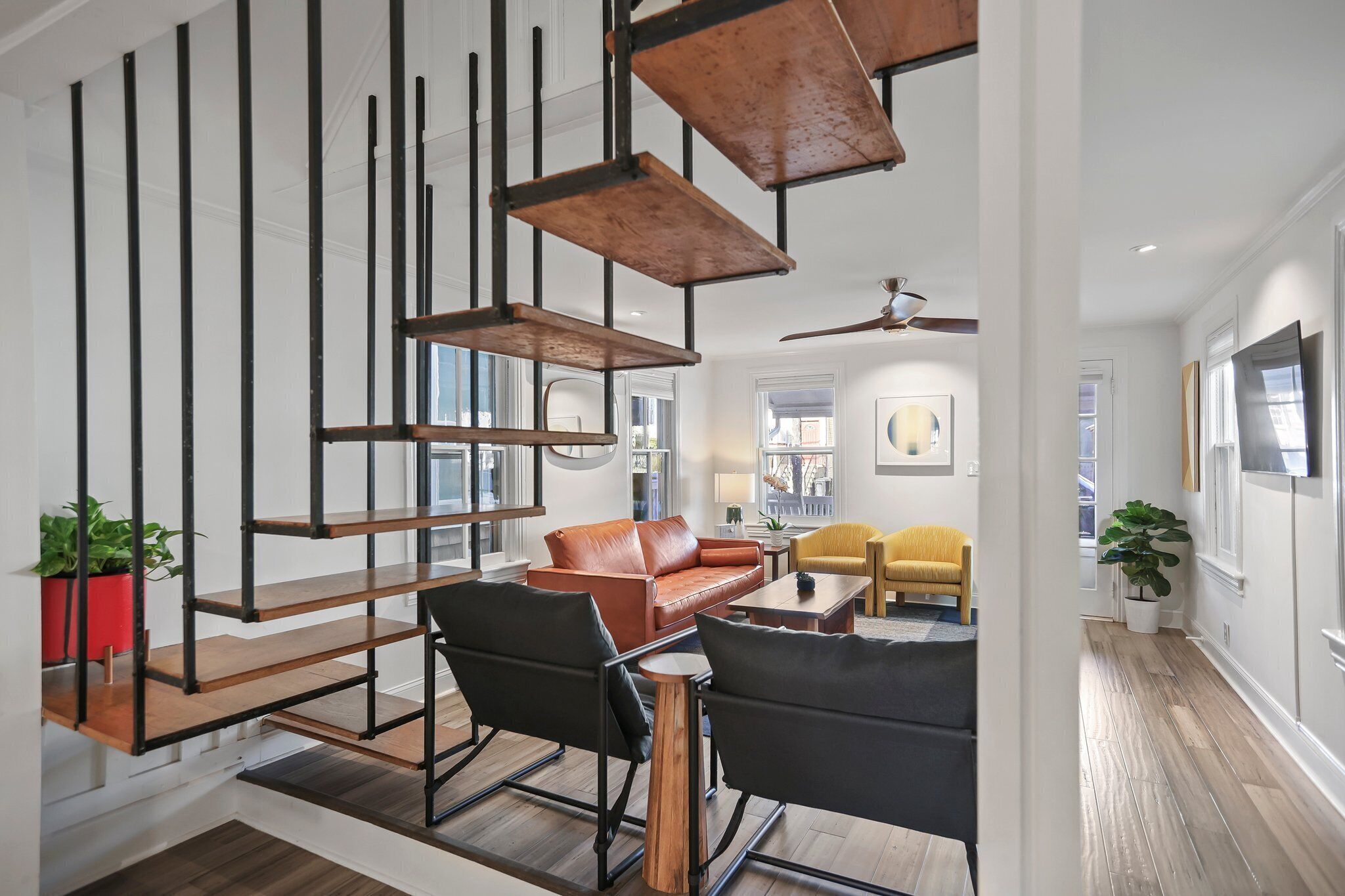 A modern living room with a brown leather sofa, black chairs, and a wooden coffee table. There are potted plants and a staircase with exposed beams.