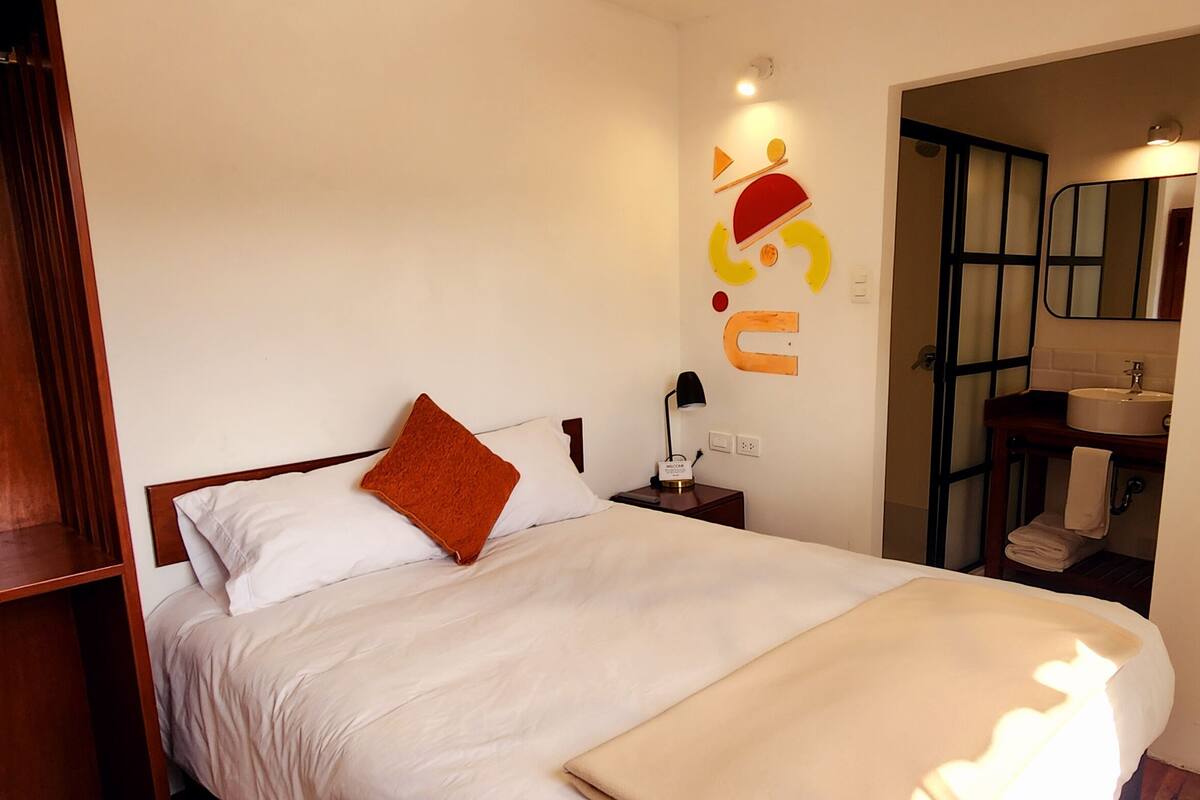Una habitación de hotel con cama, un armario de madera, una mesita de noche con lámpara y un baño visible a través de una puerta abierta.
