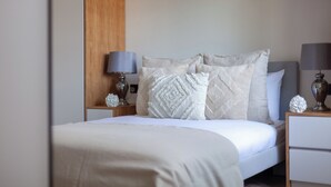 Un lit bien fait, recouvert d’une couette blanche et agrémenté de plusieurs coussins décoratifs. Il y a deux tables de chevet, chacune avec une lampe et un objet décoratif. La pièce est dotée d’un mur d’accent en bois et d’un plafond blanc uni.
