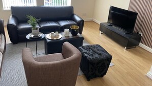 A modern living room with a black leather sofa, a coffee table with decorative items, a black ottoman, a television on a black stand, and a view of buildings through large windows. - Stylish 1-bed Apt - Sleeps 4 - Parking - Wifi (Manchester)