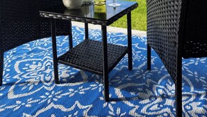A wicker table with a bottle, glass, and bowl on a patterned rug, overlooking a grassy field and hills. - Nanook’s Den Escape to the beauty of the mountains               Internet, GBTV (Petit Étang Cheticamp)