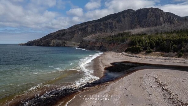 A coastal landscape with a beach, rocky cliffs, and a mountain in the background. - Nanook’s Den Escape to the beauty of the mountains               Internet, GBTV (Petit Étang Cheticamp)
