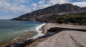 A coastal landscape with a beach, rocky cliffs, and a mountain in the background. - Nanook’s Den Escape to the beauty of the mountains, Internet, GBTV (Petit Étang Cheticamp)