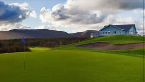 A golf course with a flagstick, a house on a hill, and mountains in the background. - Nanook’s Den Escape to the beauty of the mountains               Internet, GBTV (Petit Étang Cheticamp)