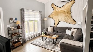A modern living room with a grey sofa, a coffee table, a mounted animal hide, and a TV. - Nanook’s Den Escape to the beauty of the mountains               Internet, GBTV (Petit Étang Cheticamp)