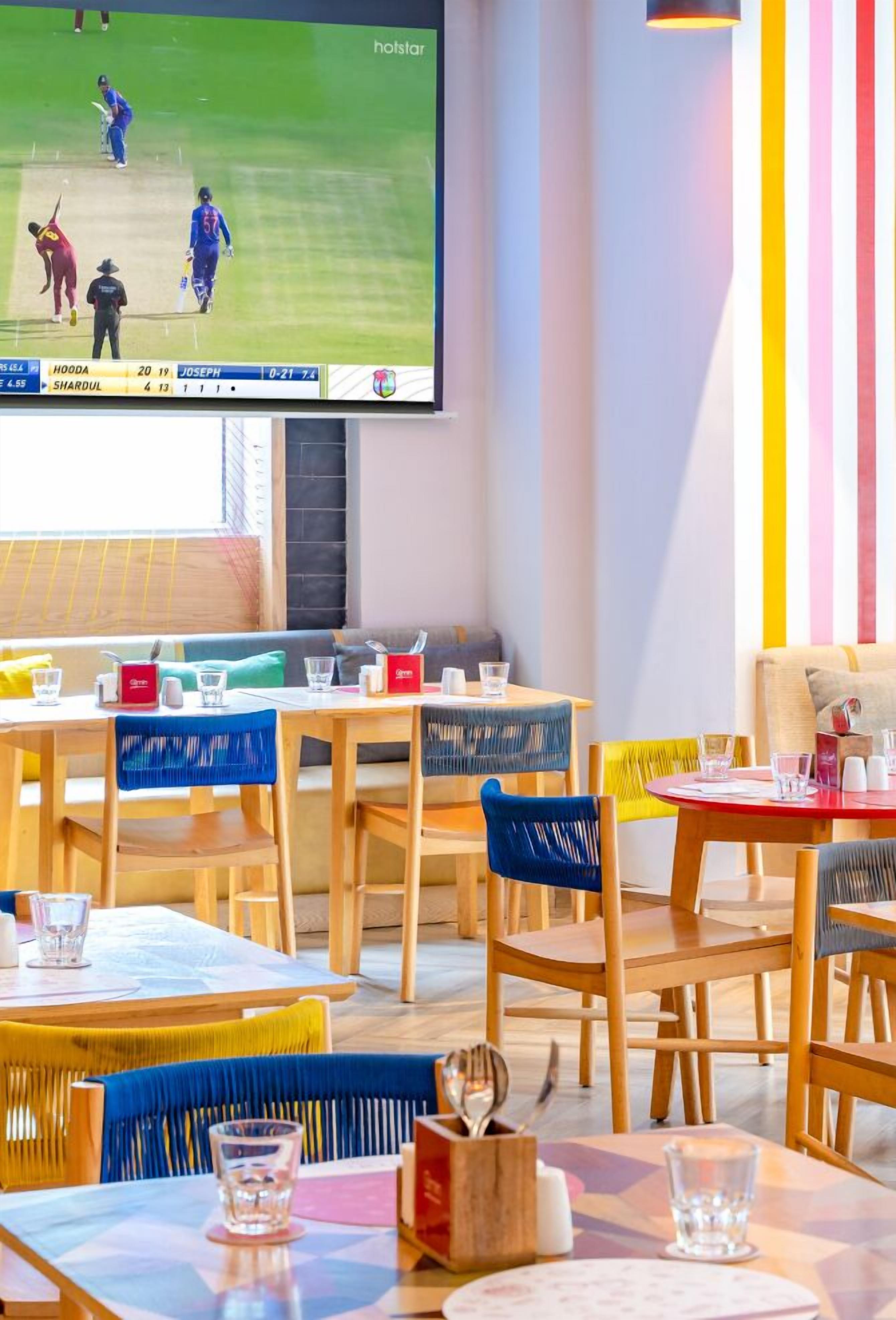 A restaurant with wooden tables and chairs, a large screen displaying a cricket match, and a colorful striped wall.