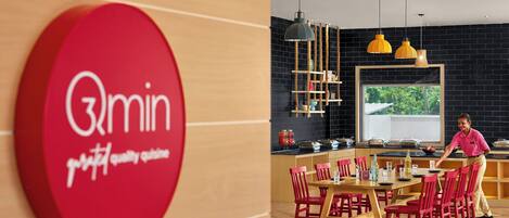 A red sign with "3min" and "quality cuisine" text, a wooden dining table with red chairs, a black brick wall, and a window with a view of greenery.