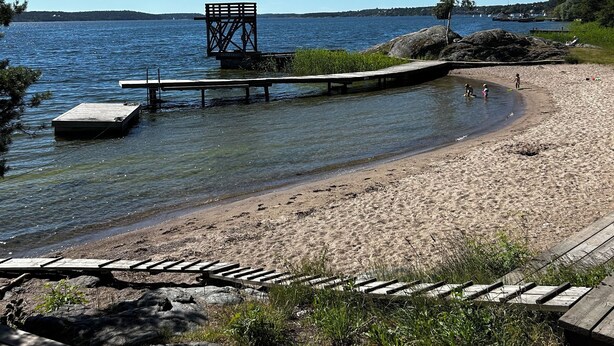 A beach with a wooden dock extending into calm waters, surrounded by greenery and a clear sky. - Cozy house with sea views, a spacious deck, and a beautiful garden (Stockholms län)
