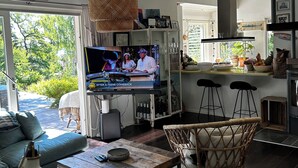 A modern living room with a sofa, a coffee table, a wicker chair, a TV, and a kitchen area with a bar. - Cozy house with sea views, a spacious deck, and a beautiful garden (Stockholms län)