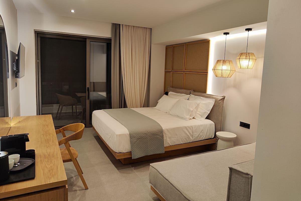 A modern hotel room with a bed, a desk with a coffee maker, a chair, and a small table.