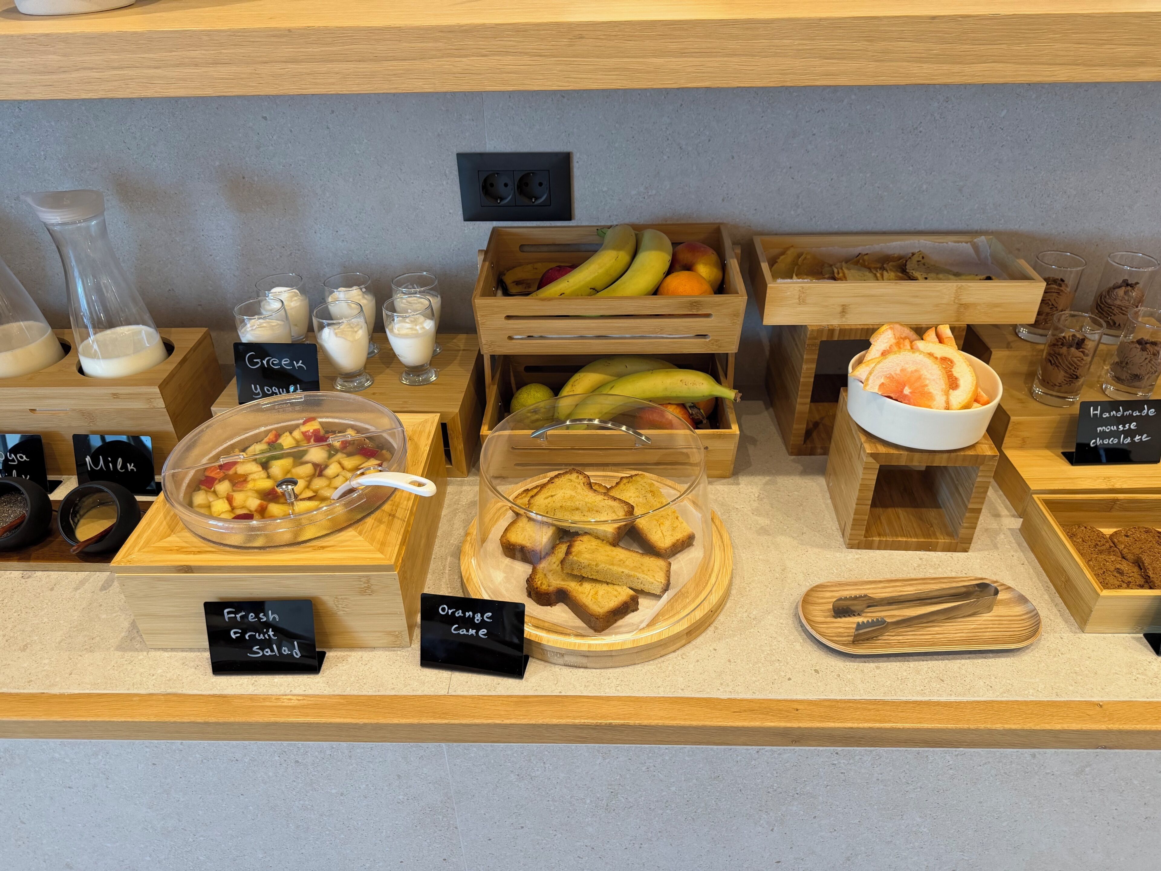 Un buffet de petit-déjeuner proposant une variété d'aliments, chacun étant clairement identifié, comme du yaourt, une salade de fruits, un gâteau à l’orange et des muffins.