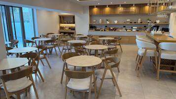 A modern cafe interior with round tables and wicker chairs, a kitchen area with wooden cabinets, and a view through large windows.