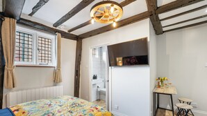 A bedroom with a bed, a window with curtains, a wooden ceiling, a small table with a vase, and a chair.