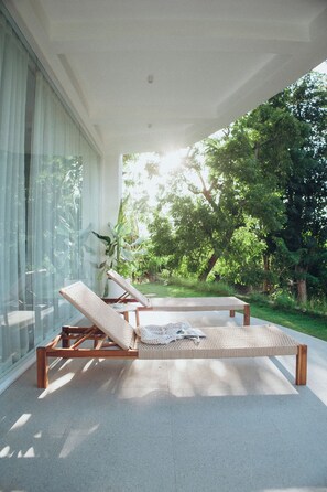 Ein moderner Außenbereich mit einer Lounge-Ecke, zwei Holzliegen und einem Tisch, umgeben von großen Glasfenstern und Blick auf einen üppigen Garten.
