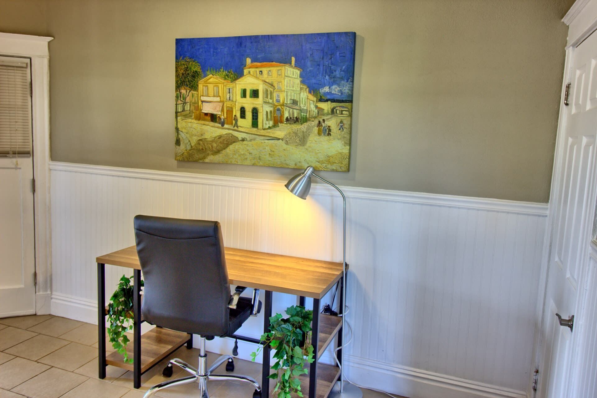 A room with a desk, chair, lamp, and a painting on the wall.
