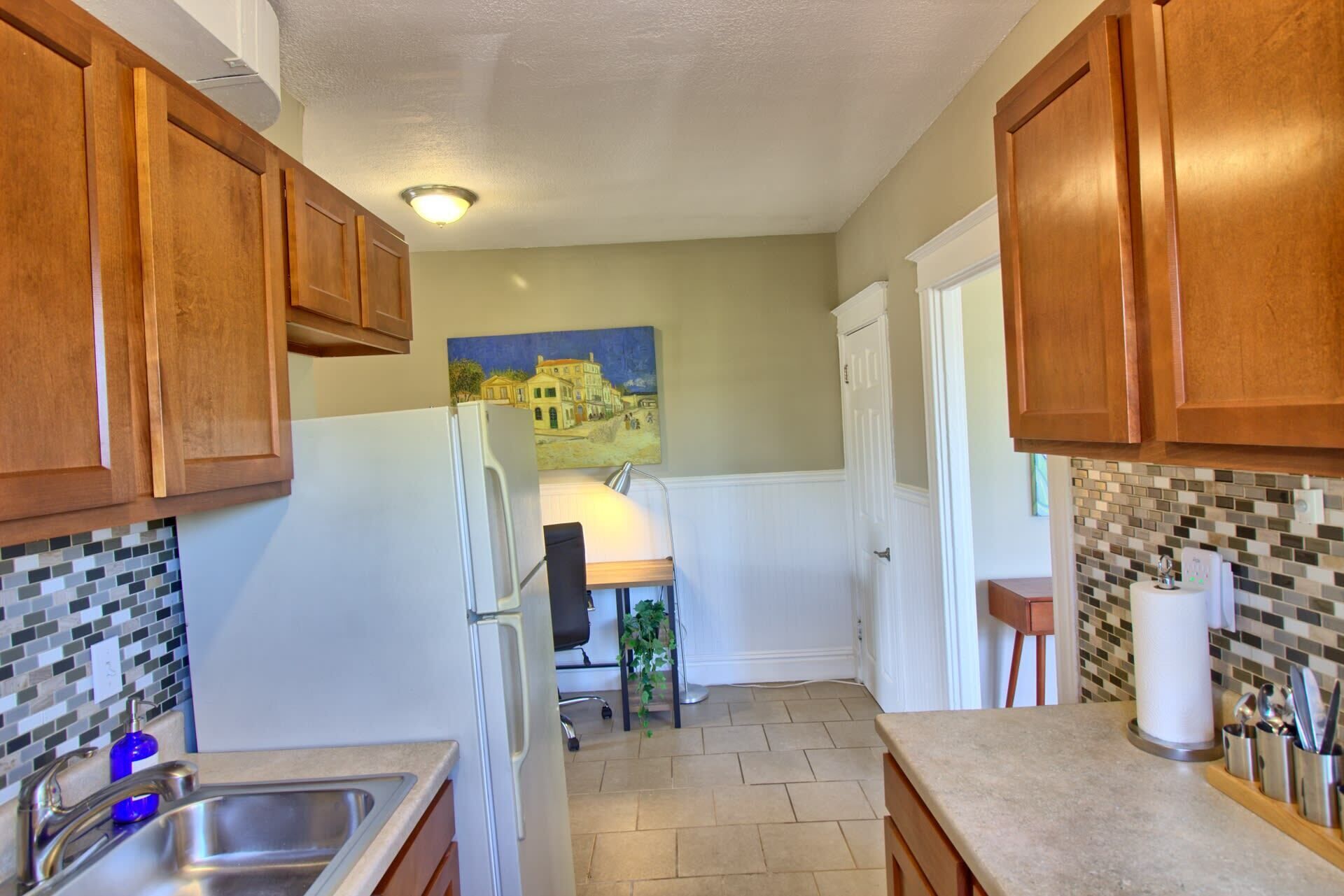 A kitchen with wooden cabinets, a white refrigerator, a tiled backsplash, and a painting on the wall.