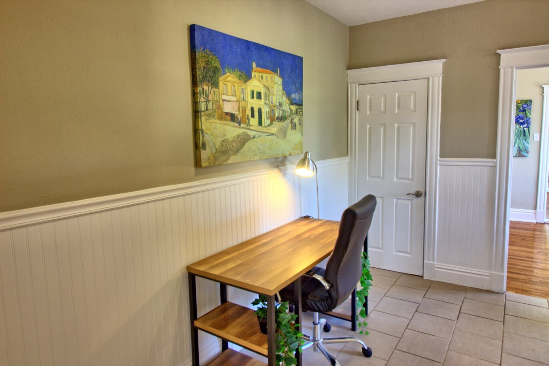A room with a wooden table, a black office chair, a white door, and a painting on the wall.